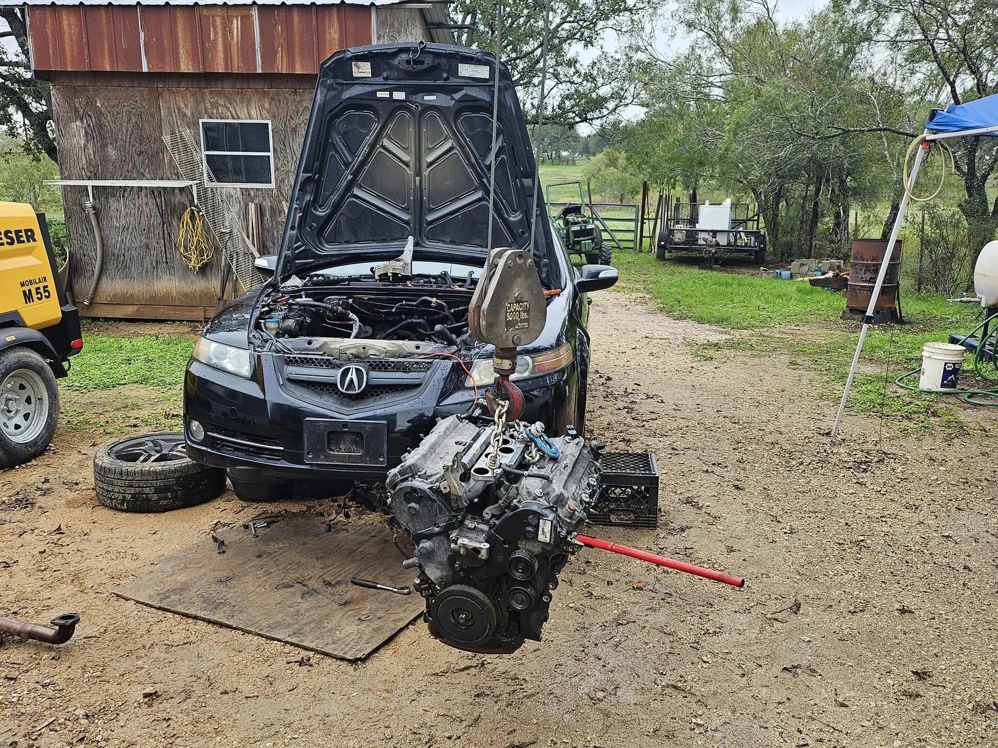 Black car with its hood open, having it entire engine remove by a crane.